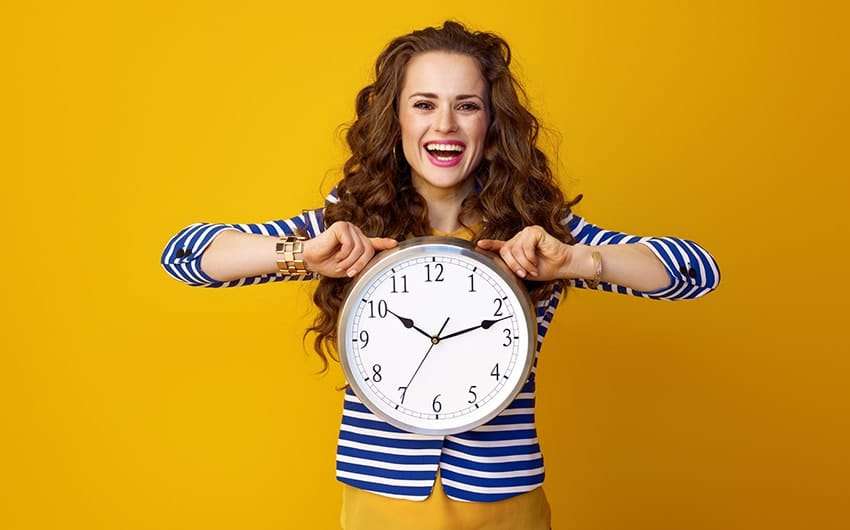 Attractive, stylish woman against a yellow background shows off her smile and a large clock. If you’re toying with the idea of braces or perhaps one of your children need braces, you might wonder when the best time to get braces is.