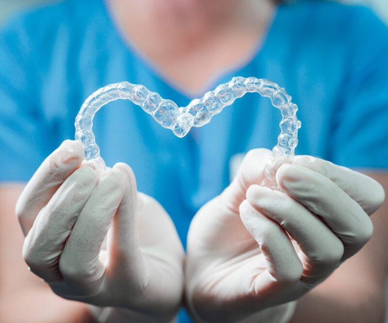 Gloved hands holding two clear Invisalign trays shaped into a heart, symbolizing comfort, care, and the personalized orthodontic experience with Invisalign