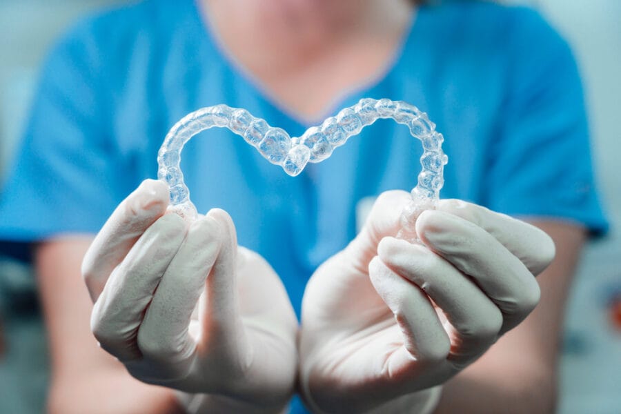 Gloved hands holding two clear Invisalign trays shaped into a heart, symbolizing comfort, care, and the personalized orthodontic experience with Invisalign