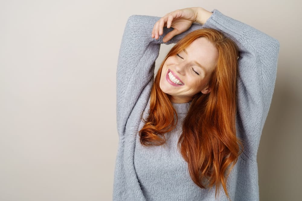 Cheerful woman with long red hair wearing a cozy gray sweater, smiling brightly with her eyes closed and arms raised - Teeth Whitening St Augustine