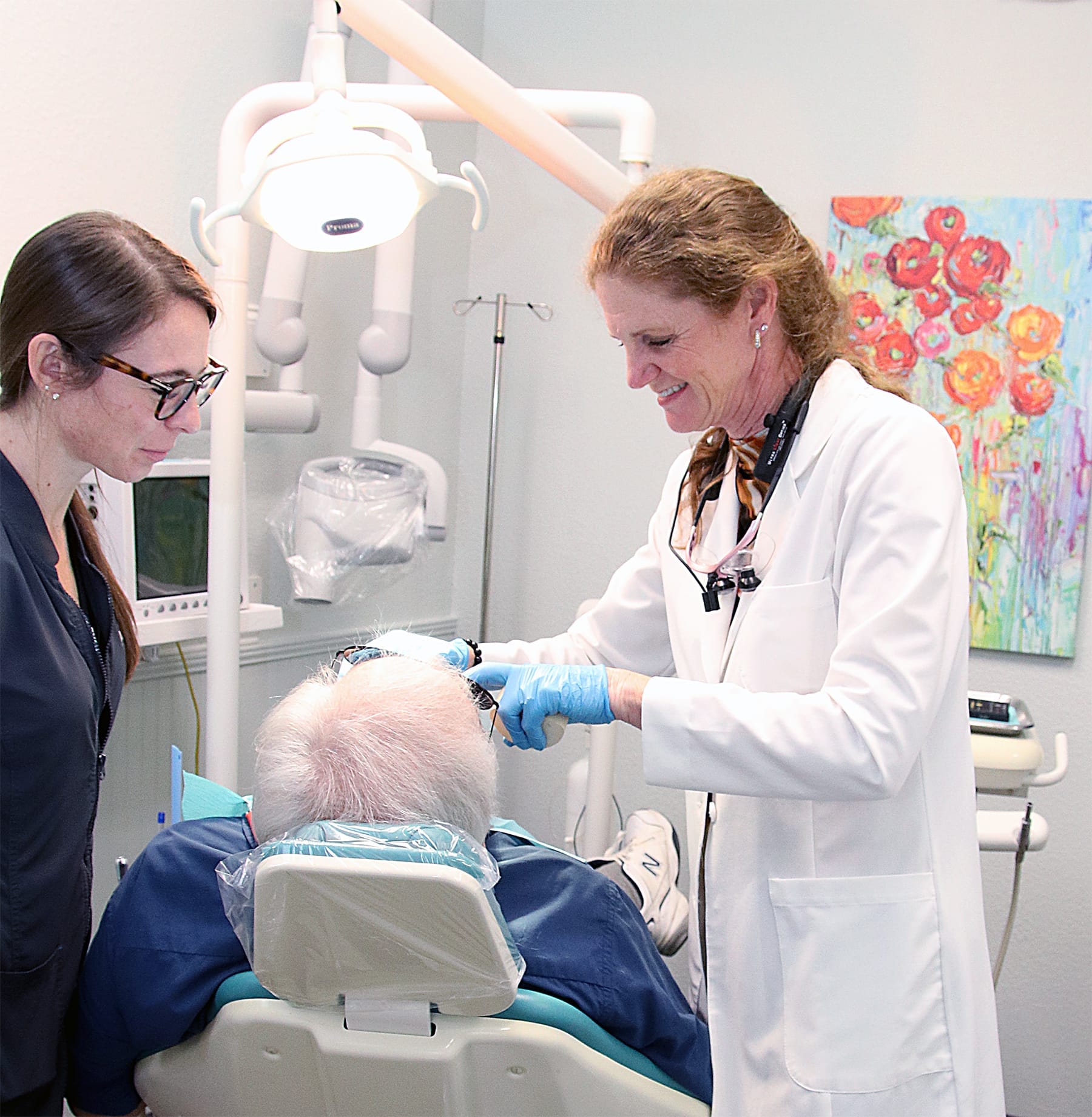 A smiling female dentist in a white coat and her assistant consulting with an elderly male patient in a dental chair, surrounded by modern equipment and a colorful flower painting – Cosmetic Dentistry