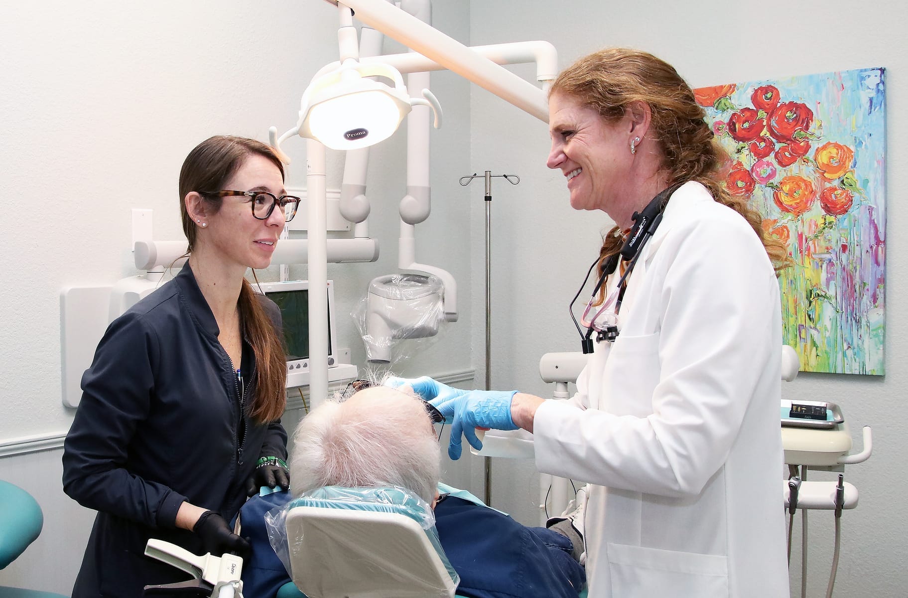 Smiling dentist and dental assistant speaking with an older male patient seated in a treatment chair, surrounded by dental equipment and colorful wall art - Dental Crowns