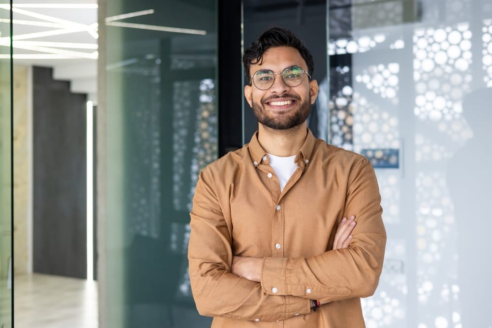 A young man wearing glasses and a brown shirt smiles confidently with his arms crossed, standing in a modern office environment – Cosmetic Dentistry St Augustine