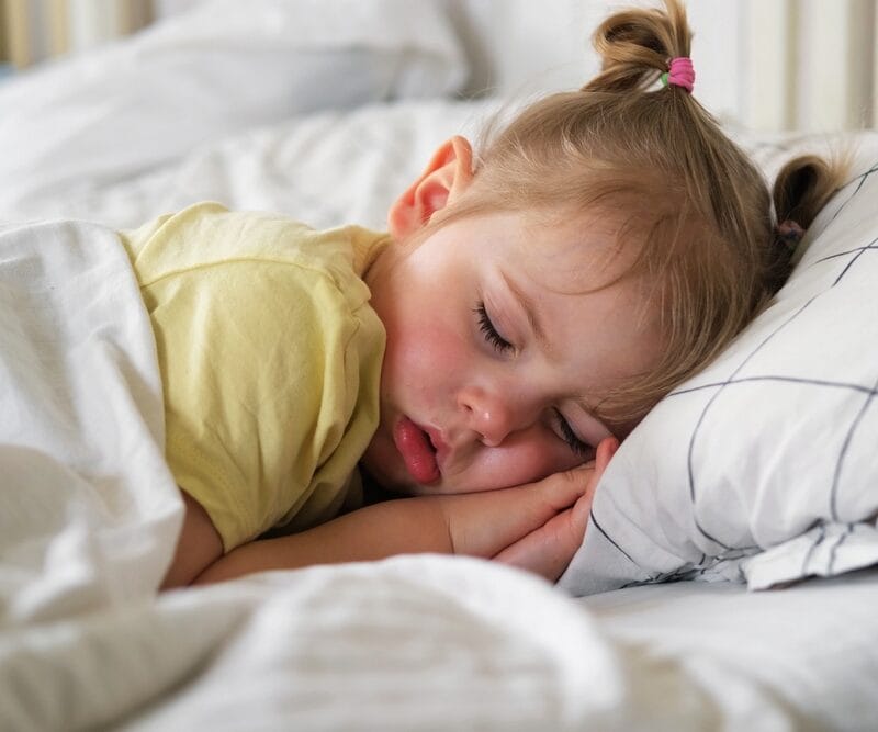 Peaceful toddler sleeping on a bed with soft white linens, representing discussions or concerns around nighttime routines when an older child wet the bed