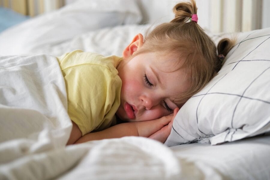 Peaceful toddler sleeping on a bed with soft white linens, representing discussions or concerns around nighttime routines when an older child wet the bed