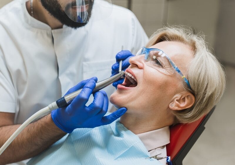 Senior woman undergoing a dental procedure while sitting in a chair, wearing protective glasses, with a dentist examining and treating missing teeth using professional dental equipment