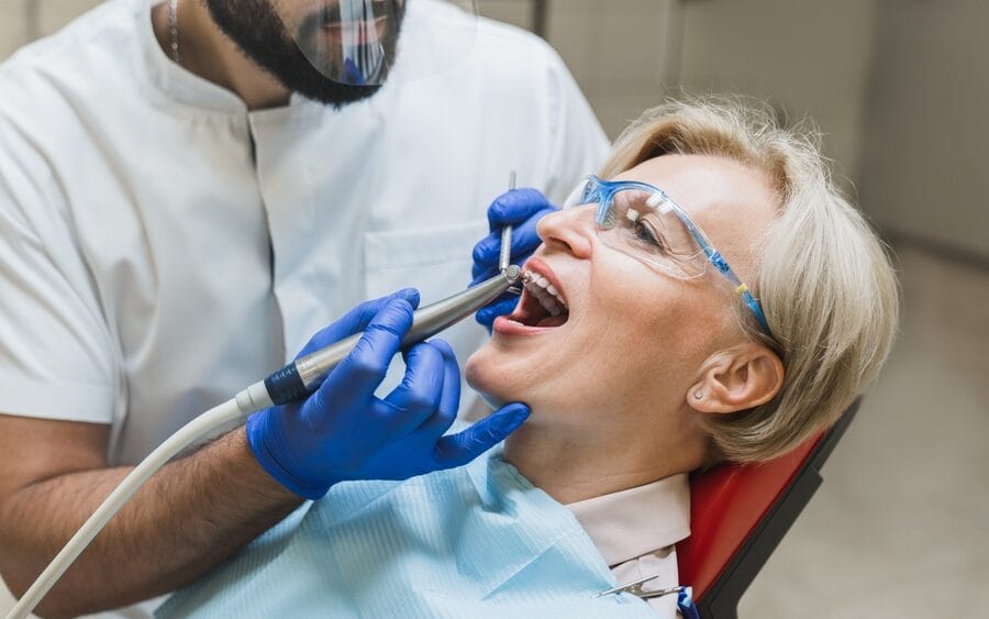 Senior woman undergoing a dental procedure while sitting in a chair, wearing protective glasses, with a dentist examining and treating missing teeth using professional dental equipment
