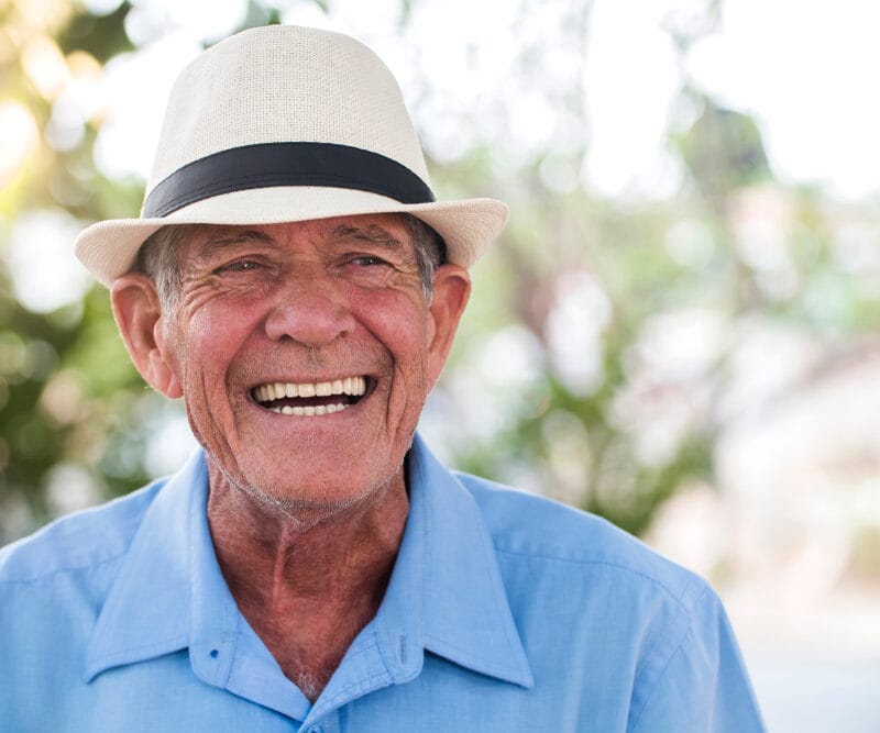 Smiling older man in a light blue shirt and fedora, exuding happiness and confidence, showcasing the emotional and aesthetic benefits of a smile makeover