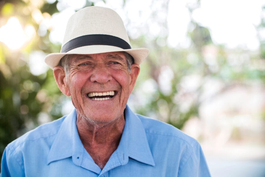 Smiling older man in a light blue shirt and fedora, exuding happiness and confidence, showcasing the emotional and aesthetic benefits of a smile makeover