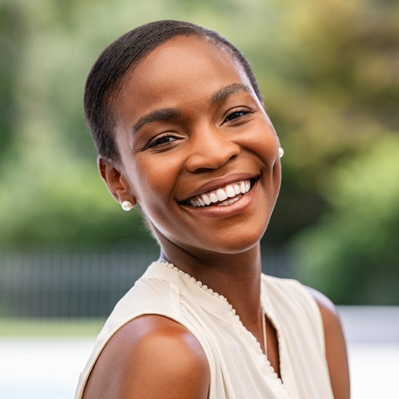 Confident woman with a bright, joyful smile against a blurred outdoor background, symbolizing timeless beauty and lasting confidence, just like the charm of St. Augustine
