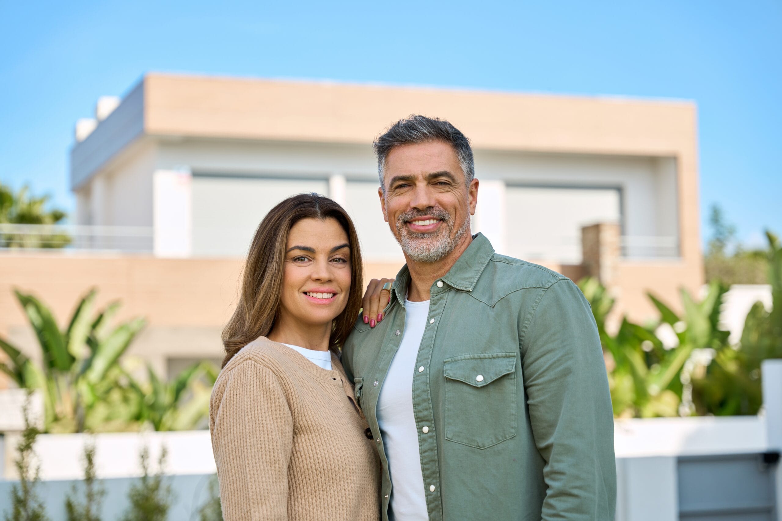 Happy middle-aged couple posing outdoors in front of a contemporary house, both smiling and dressed casually with tropical greenery in the background - Cosmetic Dentistry